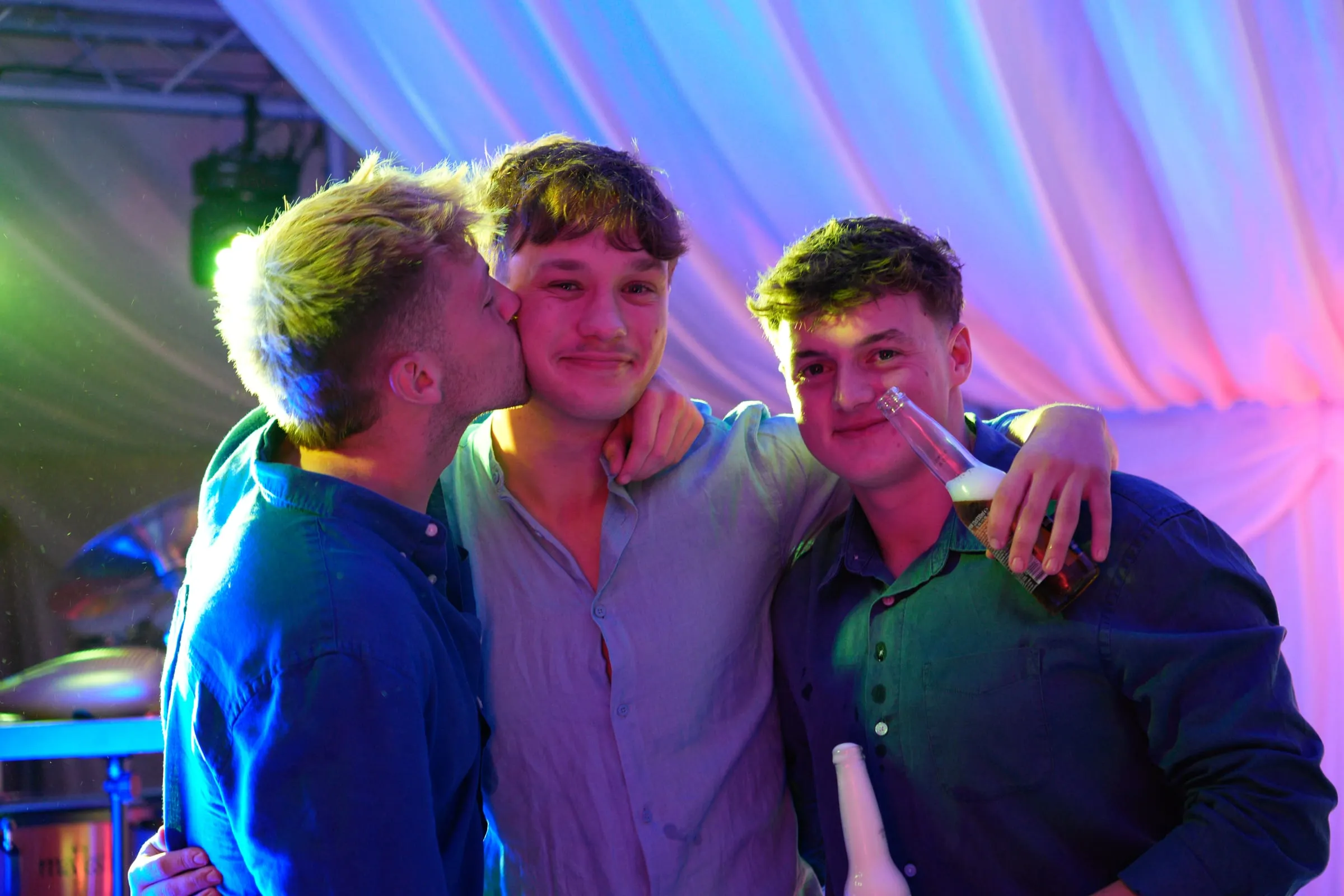 Three friends in a hug, one kissing another's cheek, under violet light.