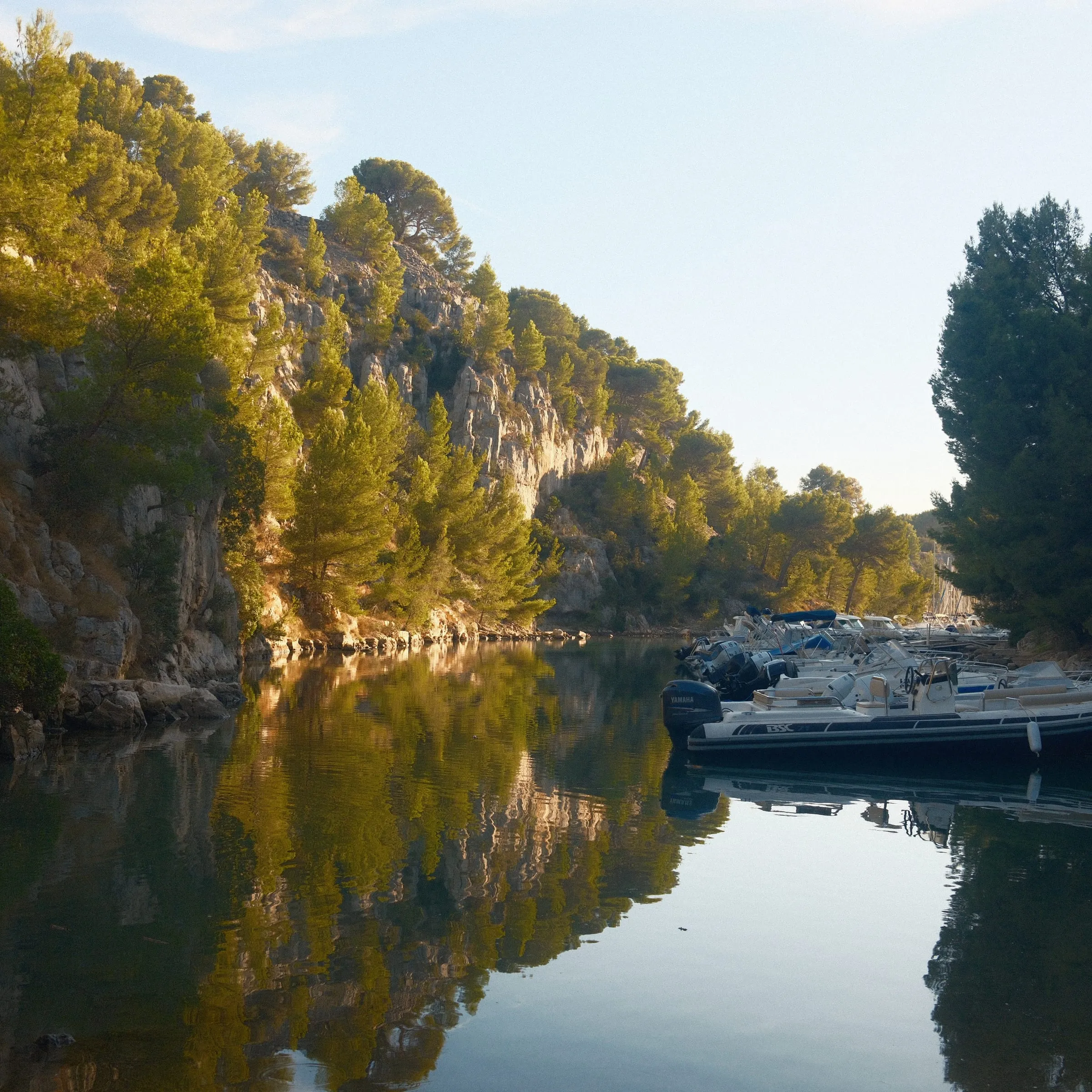 Moored boats and pine-covered cliffs reflected in still water.