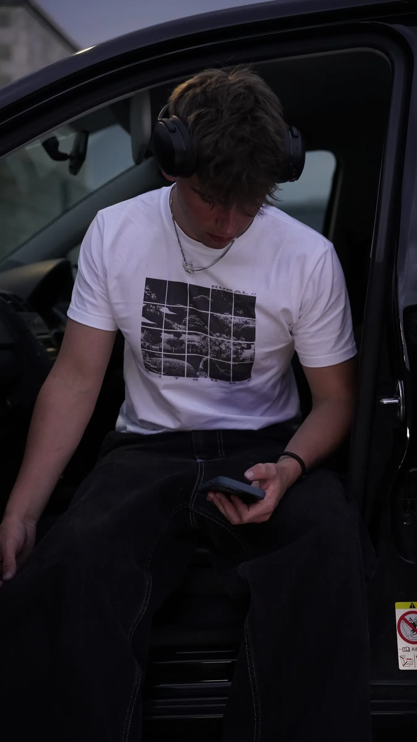 A model sits in an open car door, looking down at his phone.