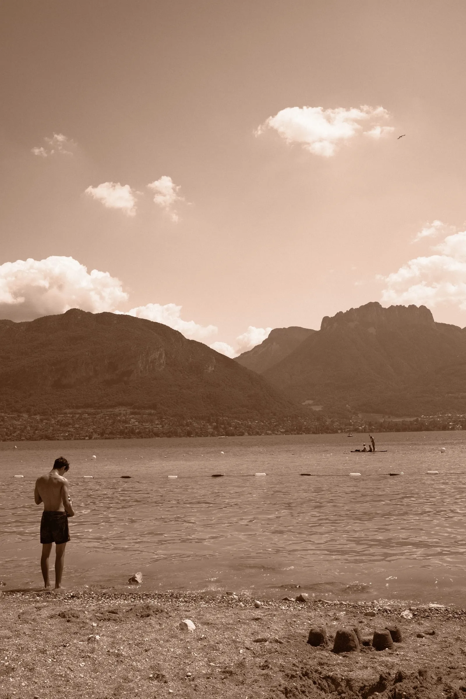 A boy stands on a pebbled lakeshore beneath the mountains, toned sepia.