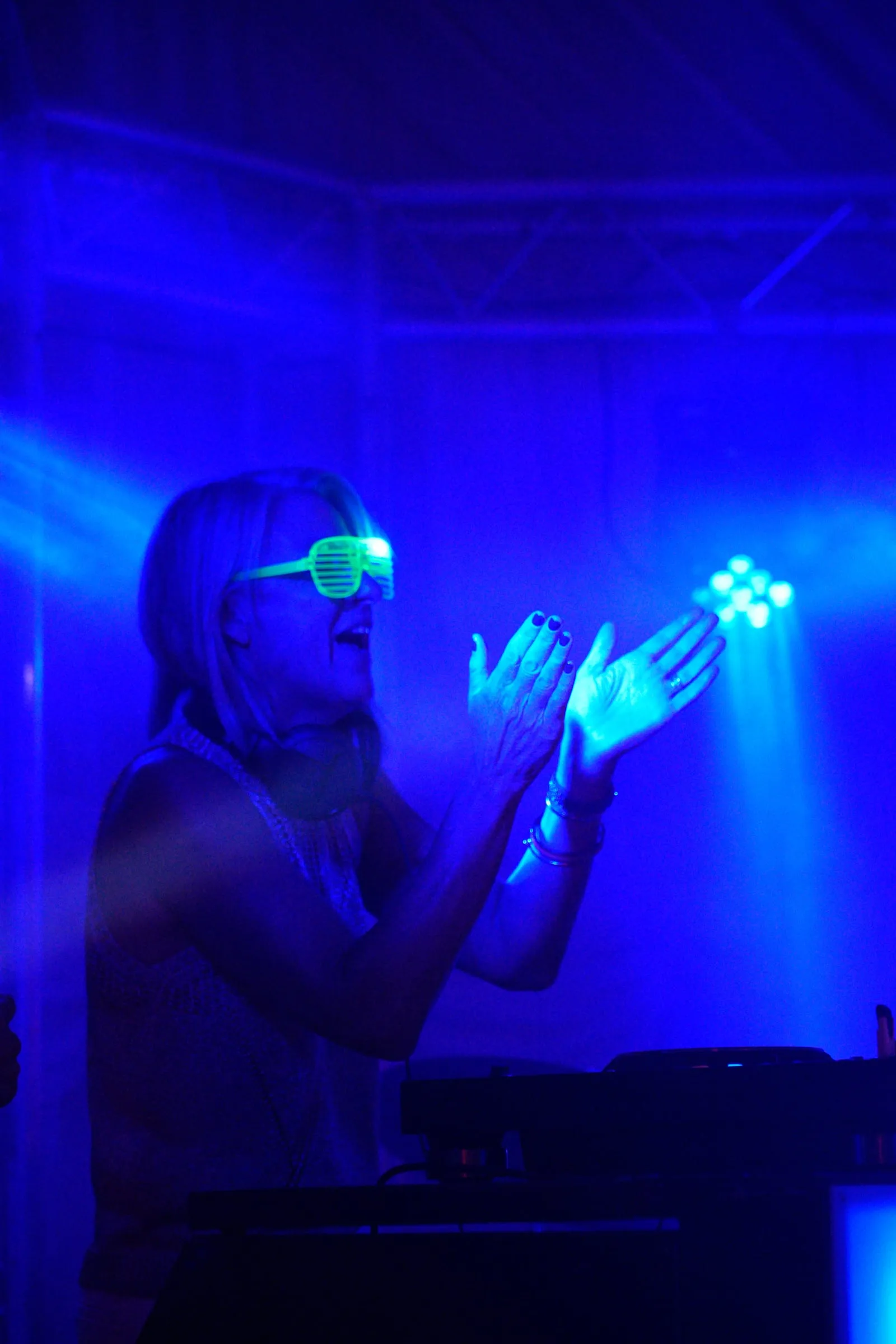 The DJ claps in glowing sunglasses, everything lit blue.