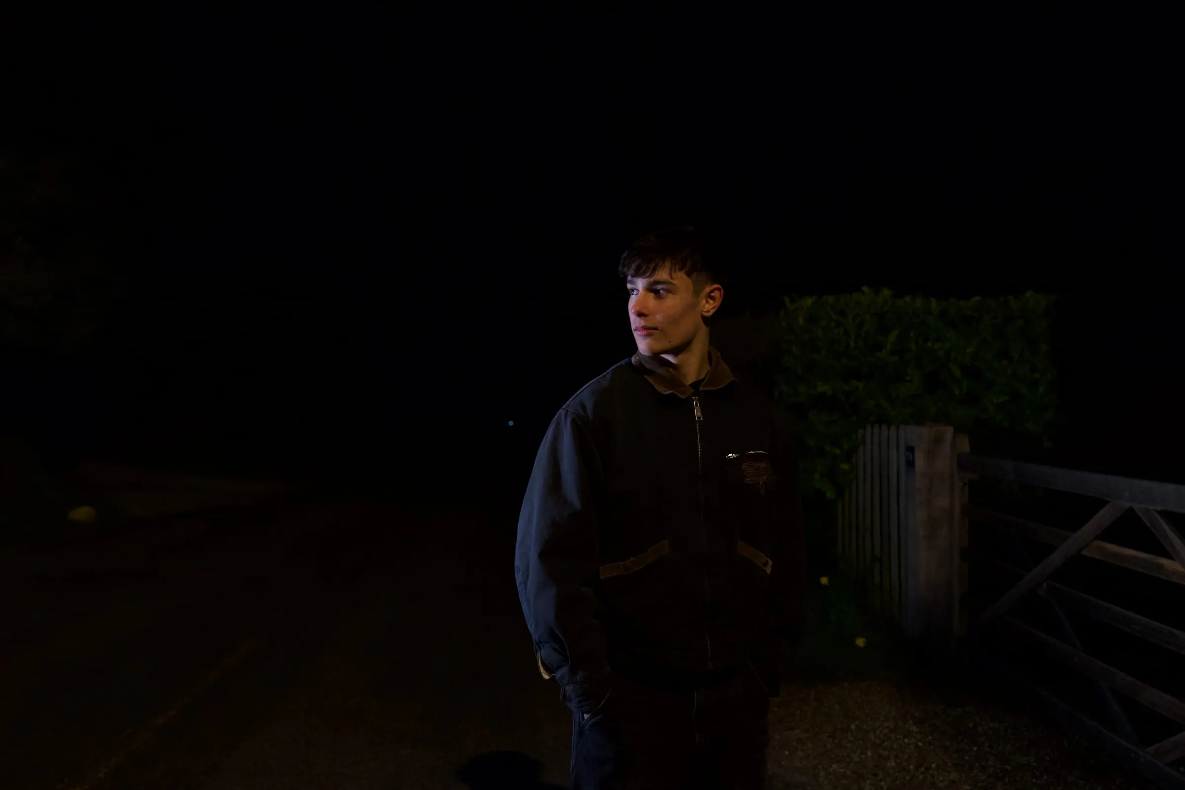 He stands by a garden gate at night, a single light on his face.